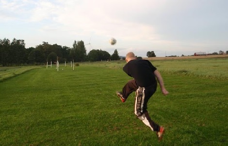 En bild på en man med svarta kläder som sparkar en fotboll ut över ett grönt fält.