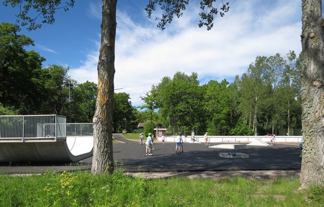En foto över Ödeshögs Skatepark. På bilden ser man en skatebana med en stor ramp till vänster. På asfalten finns flera barn som åker skateboard och kickbike. 