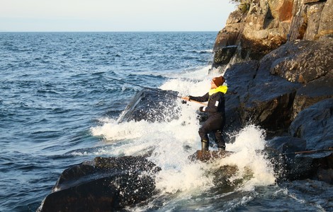 En man som står och fiskar på ett par klippor när vattnet slår mot land. Mannen har ett fiskespö pch har på sig en svart fritidsjacka, gul hoodietröja och en röd mössa.