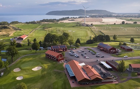 Ett flygfoto över en golf och boendeanläggning. På bilder ser man berget Omberg, sjön Vättern, stora åkerfält, golfbanor och en stor parkering med flera röda hus & byggnader som tillhör golfbanan. 