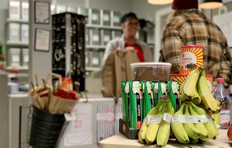 En fokusbild tagen i en butikslokal där man säljer närproducerade varor. På bilden ser man banan, russin, ost i tub och pepparkakor. I bakgrunden står en kund i rutig jacka och butikspersonal. Man kan även skymta ett smyckesställ och snittblommor. 