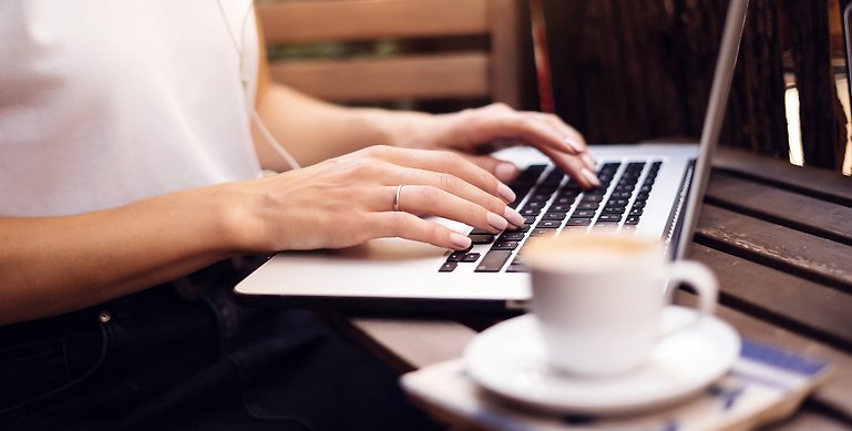 Fokusbild på en kvinna med vit t-shirt som sitter framför en laptop. Framför datorn står en vit kaffekopp. 