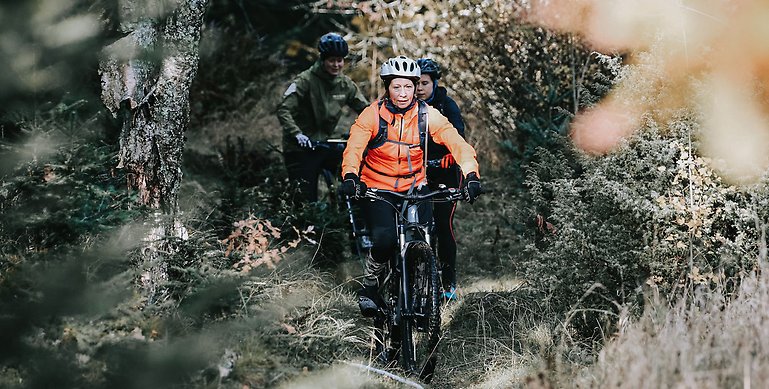 Tre cyklister som cyklar längs med en mountainbikeled i skogen. Längst fram cyklar en kvinna med orange jacka, vit hjälm och märka byxor. I bakgrunden ser man två män i mörk klädsel som cyklar strax bakom.